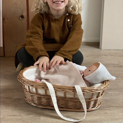 Mädchen sitzt vor dem Puppentragekorb aus Geflecht und deckt gerade die Puppe mit der weichen Decke zu. 